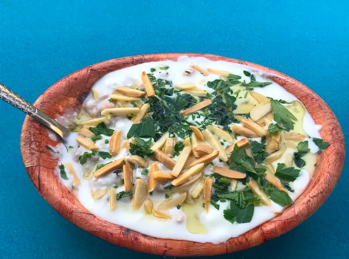 A wooden bowl brimming with yogurt, complemented by pita strips, parsley, and other toppings, set against a deep blue background