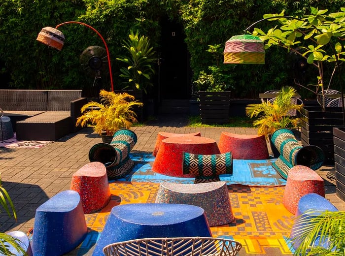 A bright outdoor patio framed by lush hedges, featuring a variety of colorful seating options arranged on a vibrant patchwork of blankets, complete with stylish arc lamps.