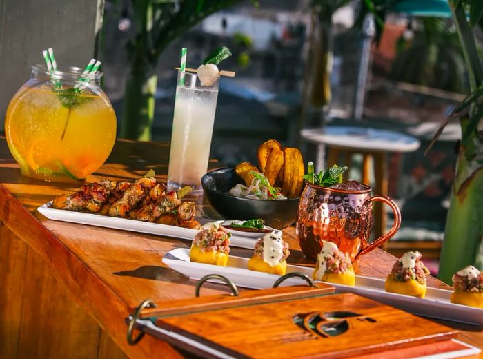 A long wooden bar is adorned with trays of assorted bar snacks, a spiral menu, an array of cocktails in various glassware, and a sizable punch bowl.