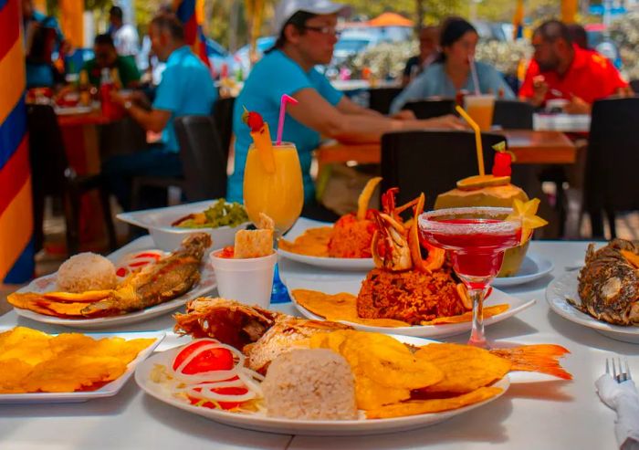 A table set outdoors is filled with dishes like fried fish, fried plantains, and cocktails, with diners in the background enjoying their meals at other outdoor tables.