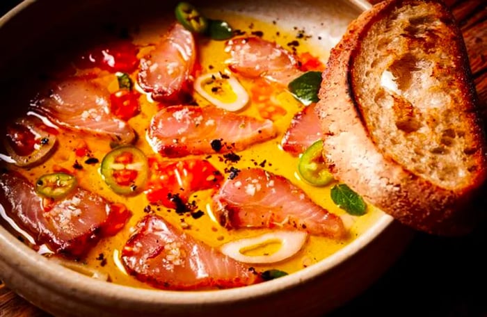 A generous bowl filled with slices of ceviche drizzled in oil, accompanied by an assortment of vegetables and a slice of bread perched on the rim.