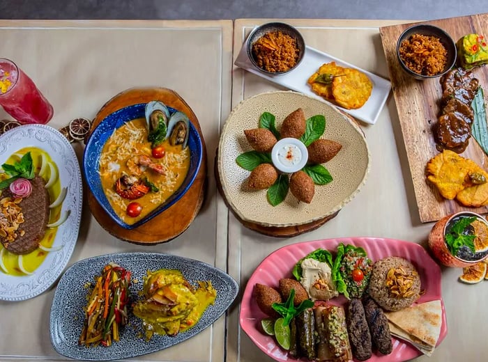 Aerial view of two small wooden tables draped with neutral-colored coverings, each adorned with an array of dishes featuring falafel, stuffed grape leaves, sirloin, fried plantains, and various dips.