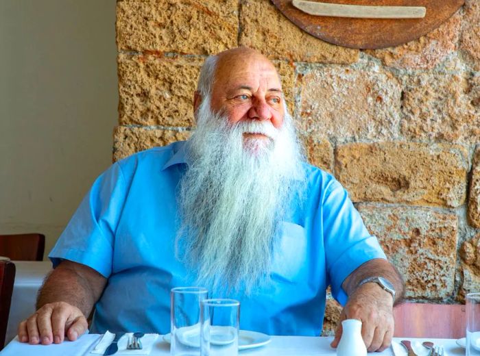 A man with a long white beard, dressed in a short-sleeved button-down shirt, sits at a beautifully arranged table against an exposed brick wall