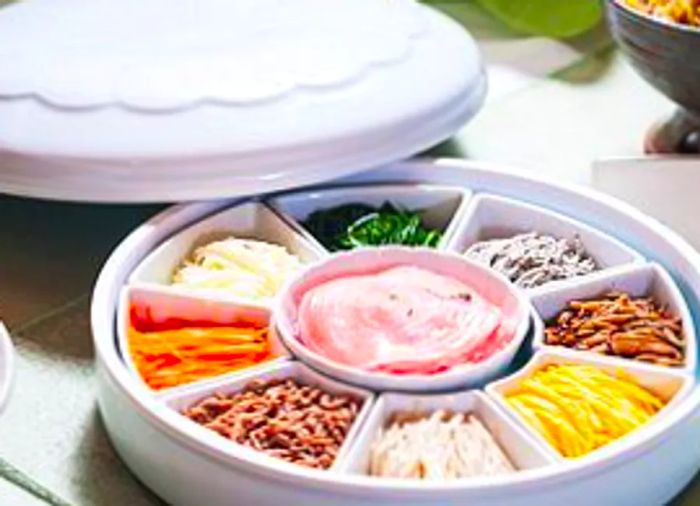 A round segmented dish featuring an array of colorful, small vegetable-based dishes in each section, surrounding a central dip, with the dish’s lid set nearby.