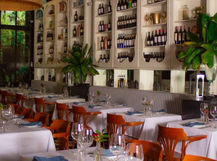 Two rows of elegantly set tables adorned with white tablecloths, one row against a tufted patterned banquette in a spacious room featuring shelves of wine on one wall and lush greenery visible through windows on the opposite wall.