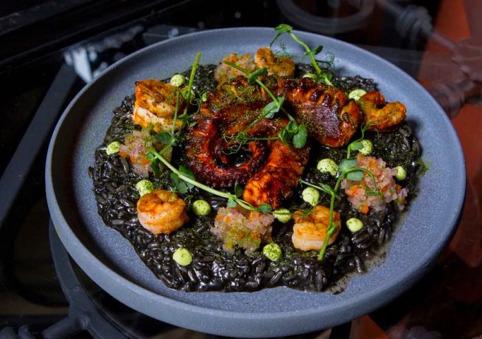 A broad, shallow dish filled with a generous portion of black rice, topped with two intertwined swirls of octopus, and surrounded by dots of assorted seafood, sauces, and fresh herbal garnishes.