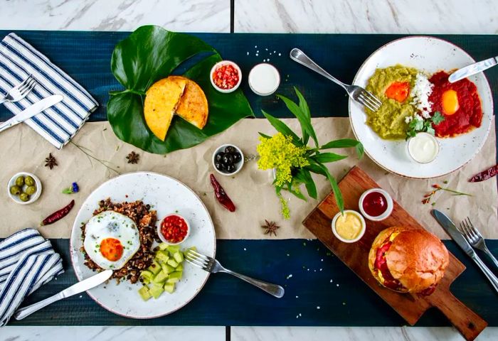 From above, a long marble table draped in blue linens features butcher paper running down the center, adorned with an array of dishes including various egg dishes, a sandwich with condiments, and a stuffed fried treat.