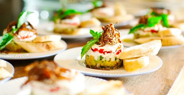 A beautifully arranged dish features layers of cheese and vegetables, topped with a small pile of fried items and a sprig of greens, accompanied by slices of bread, while several similar plates are blurred in the background.