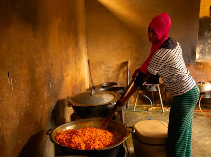 A woman stirs a large metal tray of rice alongside other cooking pots in a simple, rustic kitchen setting.