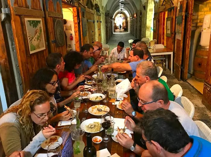 A long wooden table brimming with diners is set within a vaulted hallway, featuring heavy wooden doors lining the corridor.