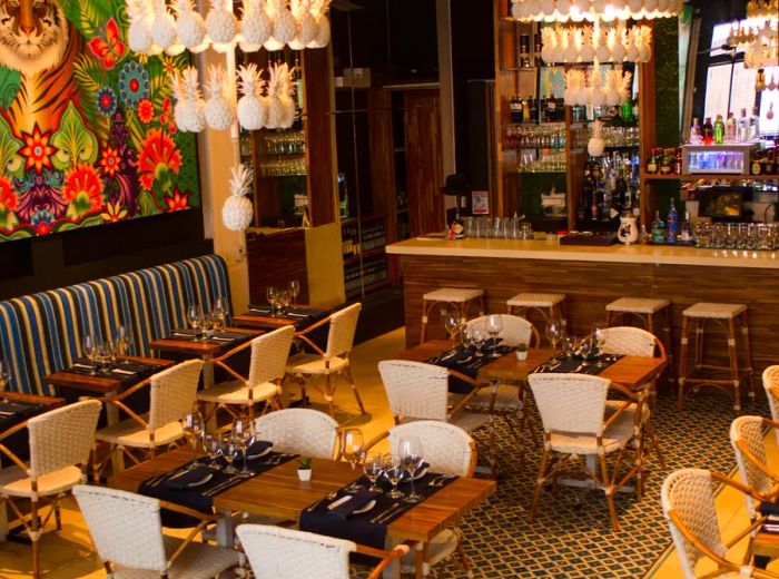 A dining area adorned with chandeliers featuring pineapple-shaped lights, a striking mural of a tiger in a jungle on one wall, wooden patio chairs with woven backs, a patterned banquette lining another wall, and a spacious bar with a mirrored backdrop.