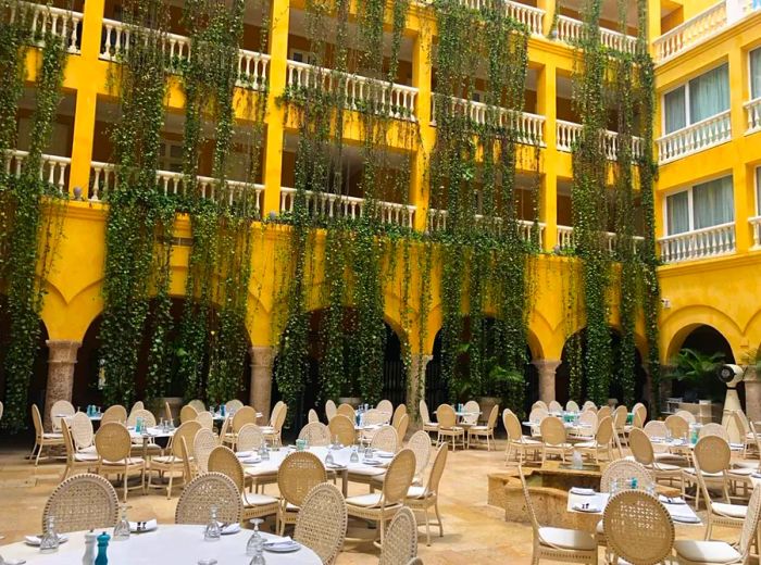 In a charming interior courtyard, spacious patio tables with woven chairs are surrounded by towering hotel buildings adorned with cascading plants that reach multiple floors.