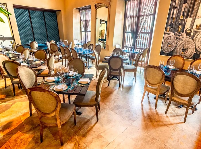 A dining room filled with natural light features rustic yet stylish tables and chairs, tiled flooring, and tall wooden doorways with windows to the outside. A striking image of ship ropes adorns the wall.