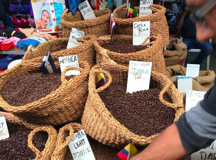 Baskets overflowing with coffee beans are adorned with handwritten signs indicating their origins, while a man reaches into the frame.