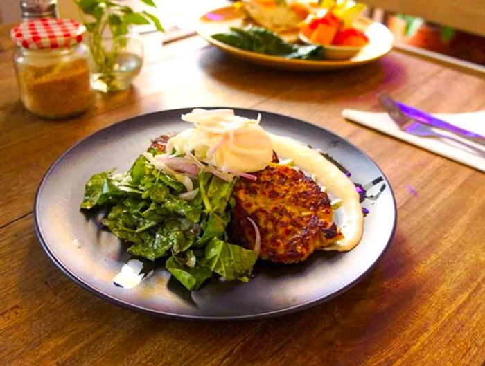 A plate showcases a mound of mixed greens next to an array of fritters, topped with a poached egg and a creamy white sauce elegantly drizzled around the dish, all set on a wooden table alongside cutlery and condiments, with another dish softly blurred in the background.