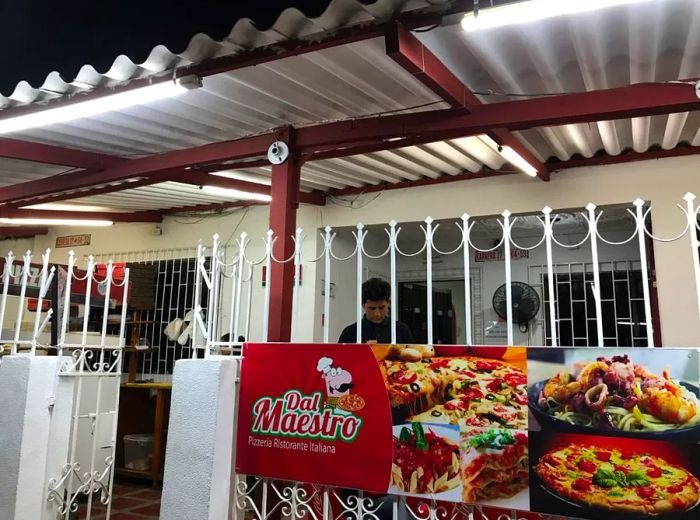 A white iron fence and gate leads to a restaurant patio shaded by a steel cover, with a poster on the fence promoting pizza and other Italian specialties.