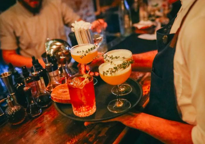 A waiter balances a tray filled with cocktails in various glass styles, some adorned with sprigs of rosemary, in front of a bar equipped with tools and ingredients, while another server is blurred in action in the background.