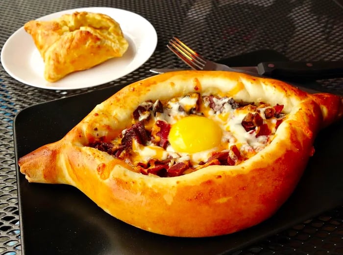 A boat-shaped Georgian <em>pide</em> flatbread filled with cheese, bits of meat, and an egg yolk at the center, presented on a large black plate, accompanied by another pastry on a separate plate, all resting on a wire mesh table.