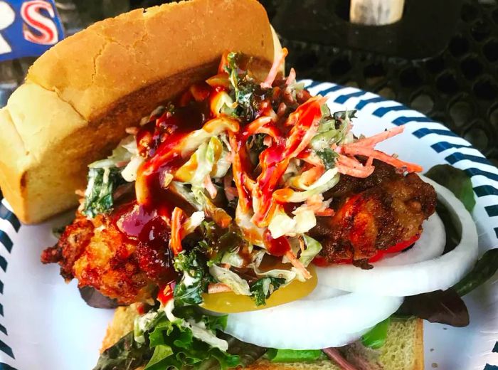 A generous pile of fried chicken sits atop a bed of slaw and onions on a slice of Texas toast, with another slice resting alongside on a striped paper plate