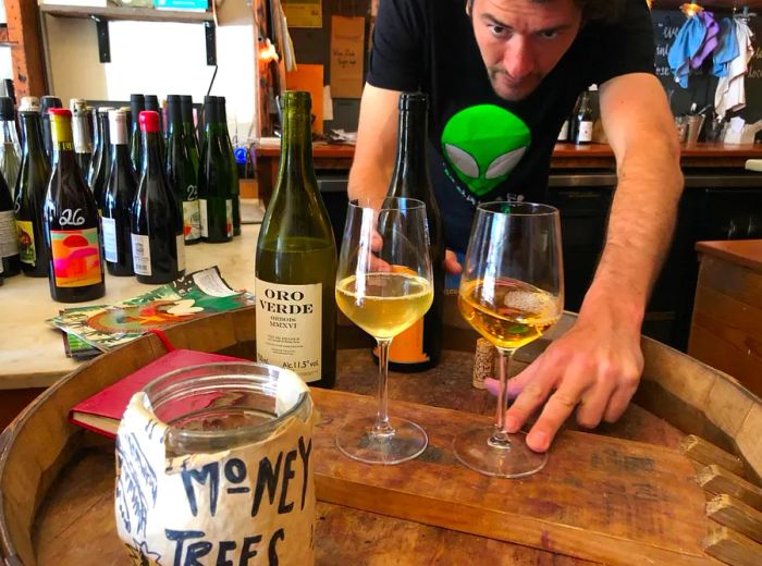 A bartender leans over a barrel-turned-table, holding two glasses of wine and an open bottle, with another table nearby filled with additional bottles and a small tip jar