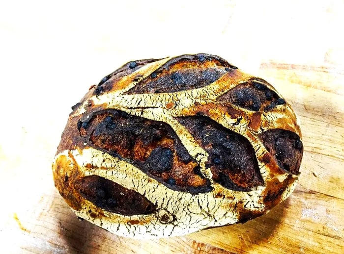 A beautiful dark loaf of bread showcasing long ripples of crust against a wooden backdrop