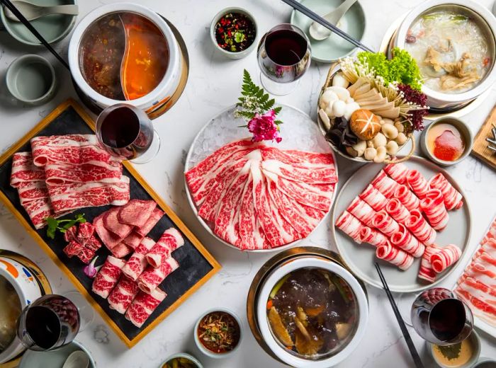 An overhead view of a table filled with large trays of assorted beef cuts, baskets of fresh vegetables, and bubbling hot pots.