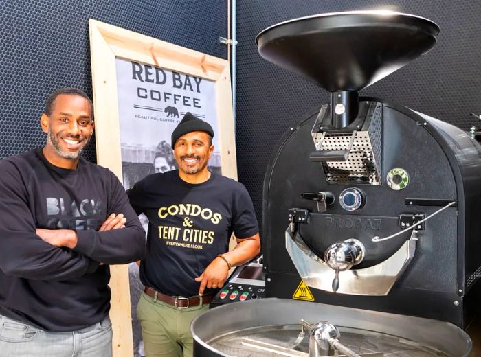 Two men stand next to a coffee roasting machine, in front of a poster showcasing Red Bay Coffee