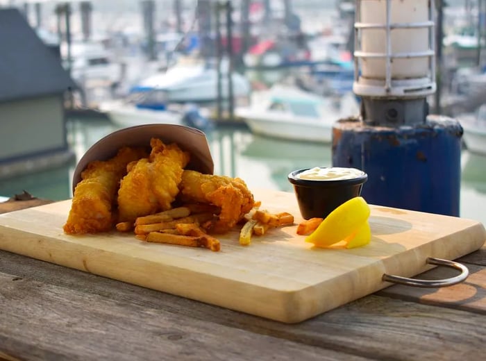 A paper cone brimming with golden fried fish and fries rests on a wooden platter, accompanied by a small cup of tartar sauce and a lemon slice, all set against a scenic waterfront backdrop.