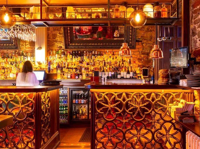 A well-lit bar featuring gold and bronze accents, an exposed stone wall adorned with large mirrors, intricate lattice bronze work beneath the counter, stylish pendant lights, and an array of bottles displayed on shelves and in fridges.