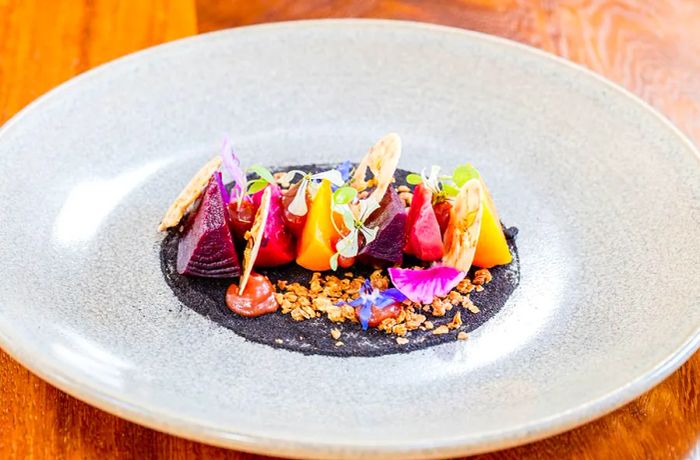 Thin slices of vibrant purple and golden beets are beautifully arranged with sprouts and a variety of colorful garnishes in a dark sauce on a pristine white plate.