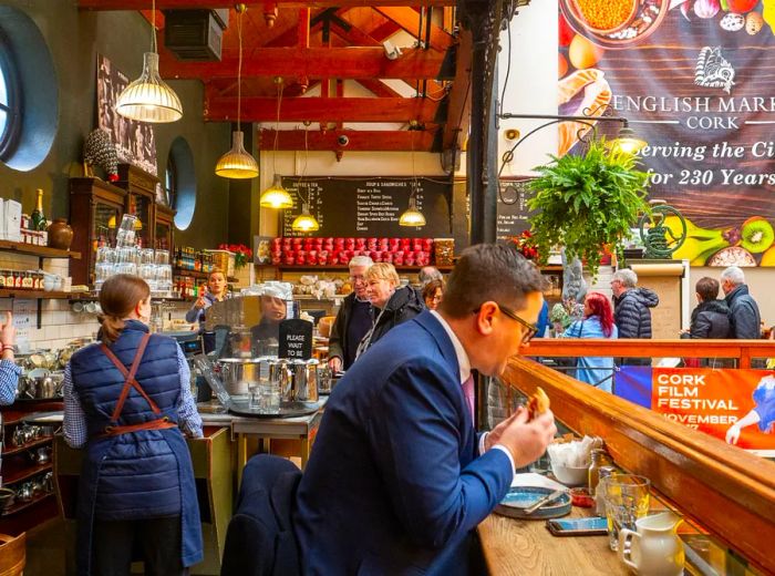 A suited man enjoys a sandwich while seated at a counter with a wooden railing, overlooking a bright atrium, as servers and patrons mingle behind him at the cafe counter and espresso preparation area.