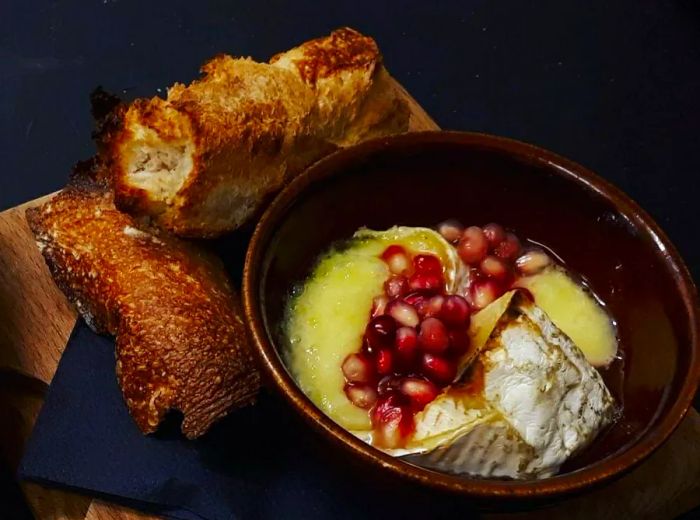 A dish featuring oozy cheese adorned with wine-soaked pomegranate seeds, accompanied by two charred slices of bread on a wooden board with a cocktail napkin.