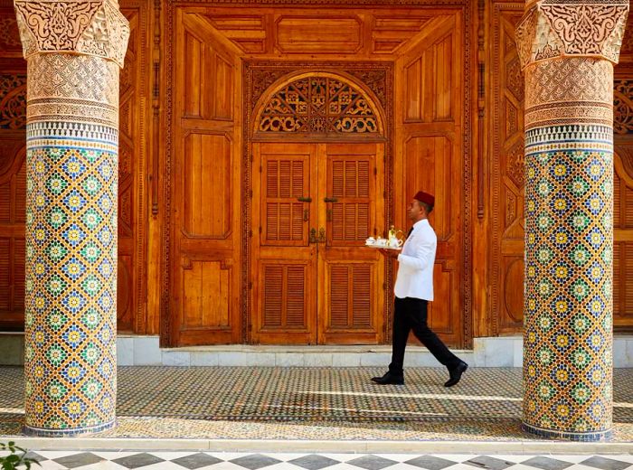 A server in a crisp white jacket and red cap carries a silver tray of tea past an impressive wall adorned with intricate woodwork, flanked by two elegant marble columns.