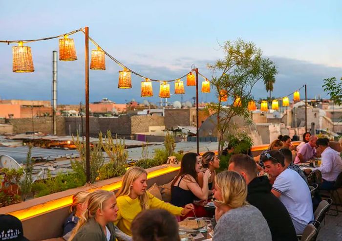 Patrons enjoy their meals at patio tables on a balcony adorned with string lights, overlooking the sprawling city of Marrakech.