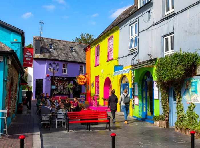 Colorful shops and buildings frame an outdoor patio on cobblestone streets, where groups of customers enjoy their time while others stroll beneath a brilliant blue sky.