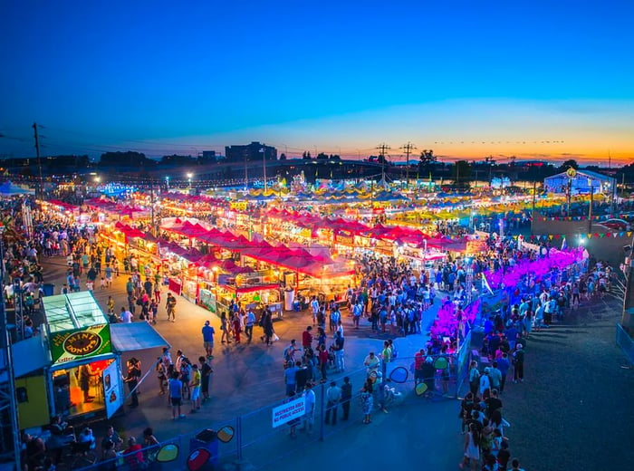 An aerial view of the night market at dusk, showcasing stalls and tents bustling with crowds lining up and browsing the stands, with a large stage at one end and the cityscape of Richmond in the background.