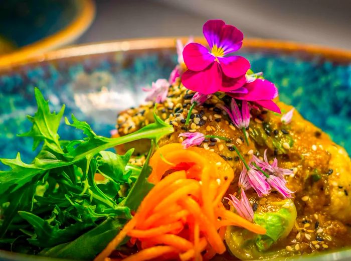 A vibrant, glossy bowl filled with an assortment of mashed and sliced vegetables is topped with a stunning floral garnish.