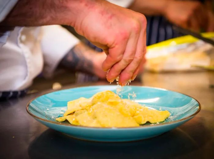 A chef adorns large pasta pieces with a sprinkle of cheese in a vibrant ceramic bowl set on a kitchen counter.