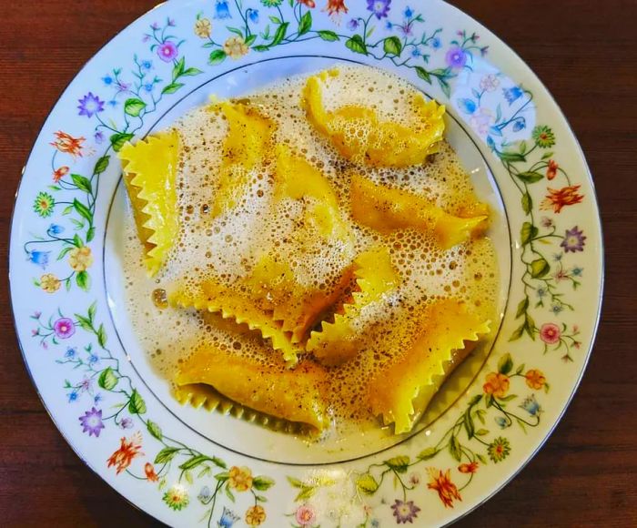 An overhead view of a floral plate showcasing a mound of agnolotti adorned with a foamy sauce