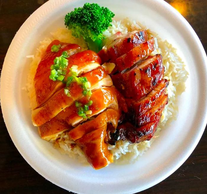 An overhead view shows sliced chicken and pork, both glistening with sauces, served over rice on a paper plate, garnished with broccoli and diced vegetables.