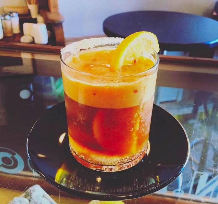 A frothy glass of espresso tonic, topped with a thick foam and garnished with an orange slice, rests on a glossy saucer atop a glass counter.