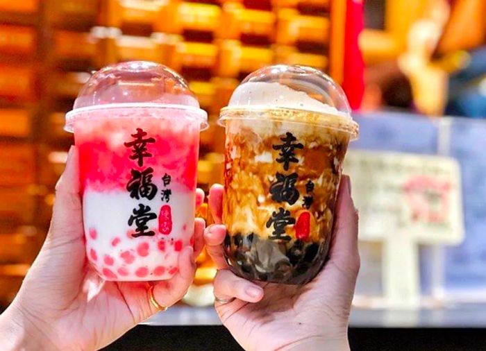 Two hands proudly display plastic cups filled with vibrant bubble tea; one cup features a layered fruit blend, while the other is swirled with rich brown sugar syrup.