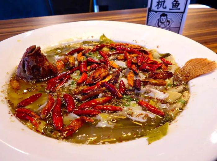 A bowl of soup brimming with chunks of fish, fiery chiles, Sichuan peppercorns, and a large fish head elegantly poised toward the sky.