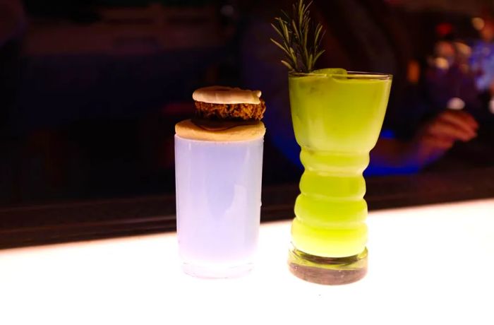 Two vibrant cocktails in decorative glassware, one topped with a cookie sandwich, on a bright, illuminated bar