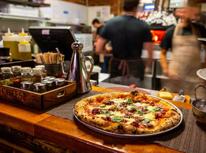 A whole pizza rests on a serving platter on a wooden counter, next to a tray filled with spice jars and an elegant olive oil dispenser, with a blurred kitchen scene and staff busy at work in the background.