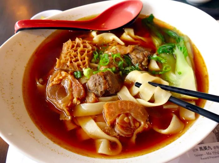 An overhead view of chopsticks lifting noodles from a bowl of soup that includes large pieces of tripe and beef shank, bok choy, and diced scallions, with a soup spoon resting at the edge of the bowl.