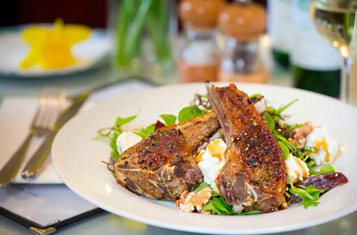 Two sizable lamb chops stand tall on a bed of greens, complemented by various accompaniments on a dinner plate next to neatly arranged cutlery and table decor.