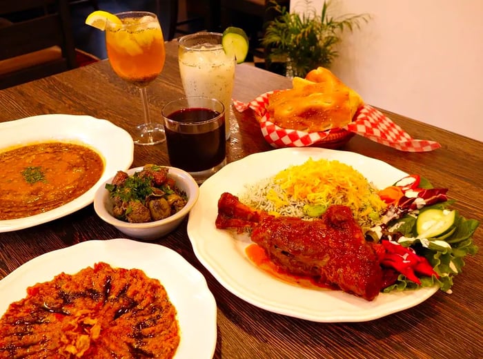 A small wooden table is filled with an array of dishes and drinks, showcasing lamb shank, a basket of bread, two dips, and cocktails alongside wine in various glasses.