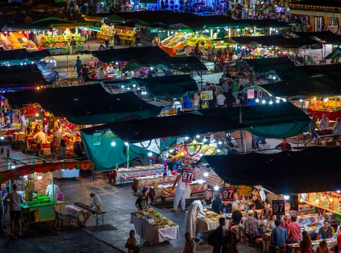 From a nearby rooftop, the Jemma el Fna market at night is alive with rows of stalls illuminated by bright lights, as vendors and customers mingle beneath awnings marked with numbers.