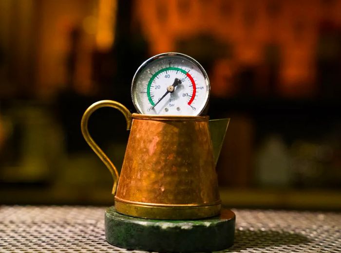 A copper saucer rests on a coaster, featuring a pressure gauge balanced on the rim, all set within a dimly lit bar atmosphere.